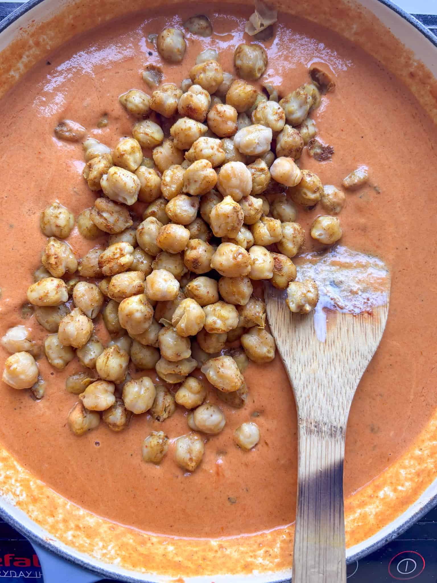 a pan of creamy tikka masala sauce with seaoned chickpeas just added to it. A spoon sits to one side ready to stir them in.
