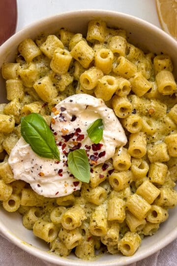 a bowl of creamy pesto ditalini pasta topped with creamy burrata, chili flakes, and basil