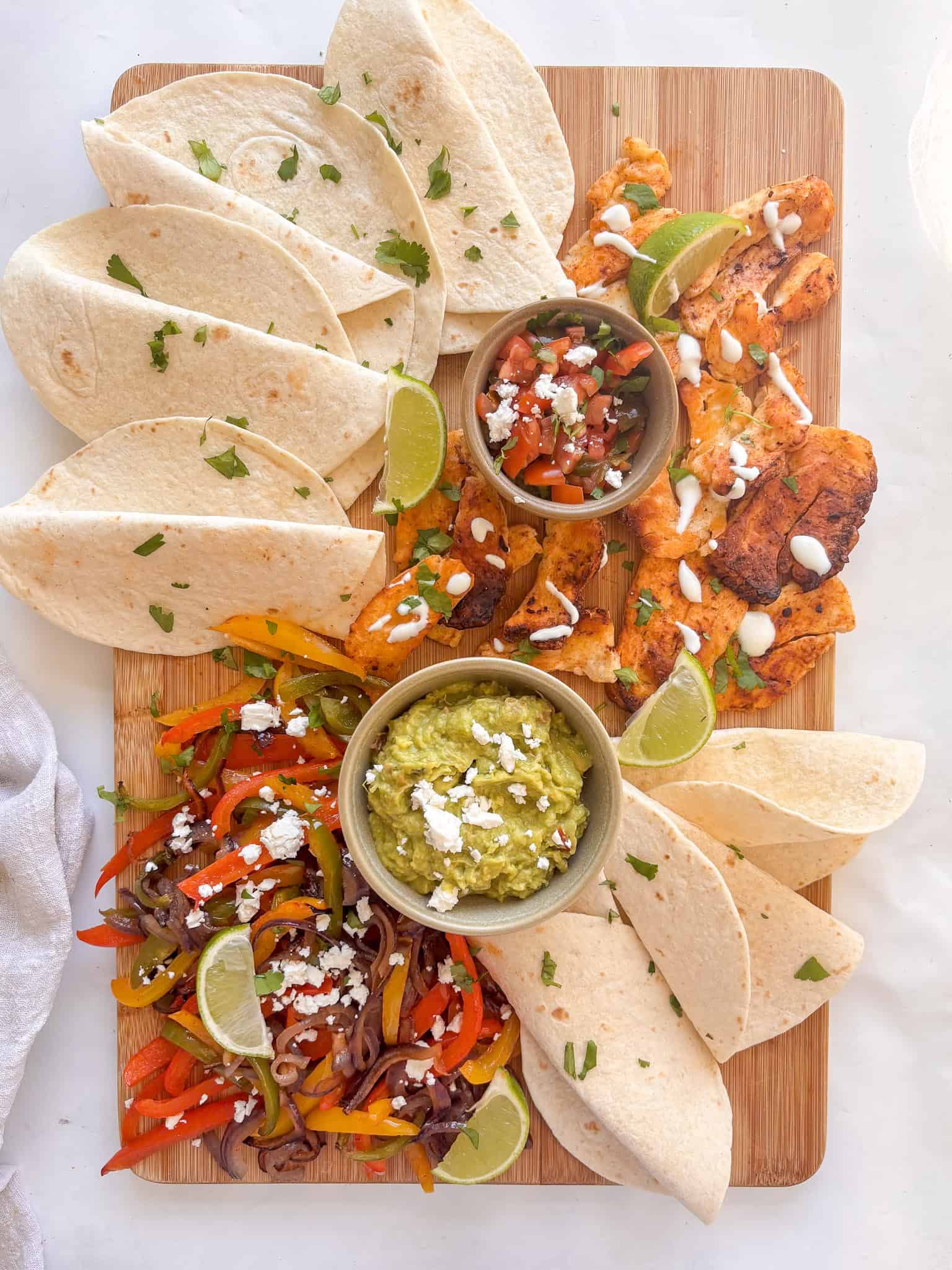 roasted halloumi and vegetable traybake on a chopping board with tortilla wraps, pice de gallo in a bowl, guacamole in a bowl, and lime wedges scattered throughout