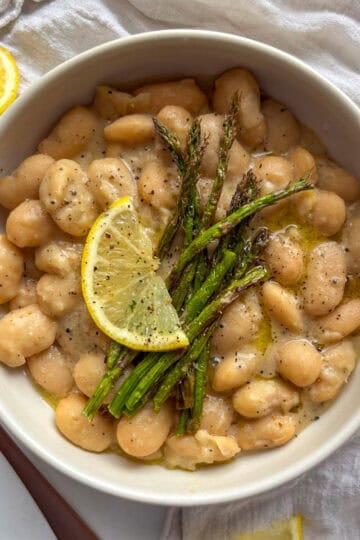 a bowl of creamy cacio e pepe beans topped with grilled asparagus and a slice of lemon